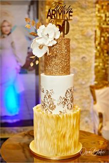 Three-tiered gold and white birthday cake with floral and sequin details, topped with a name and age (50).