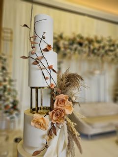 Three-tiered white wedding cake with gold accents and peach-toned floral decorations, in front of a blurred wedding backdrop.
