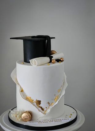 A two-tiered graduation cake. White frosting with gold accents and a black graduation cap.