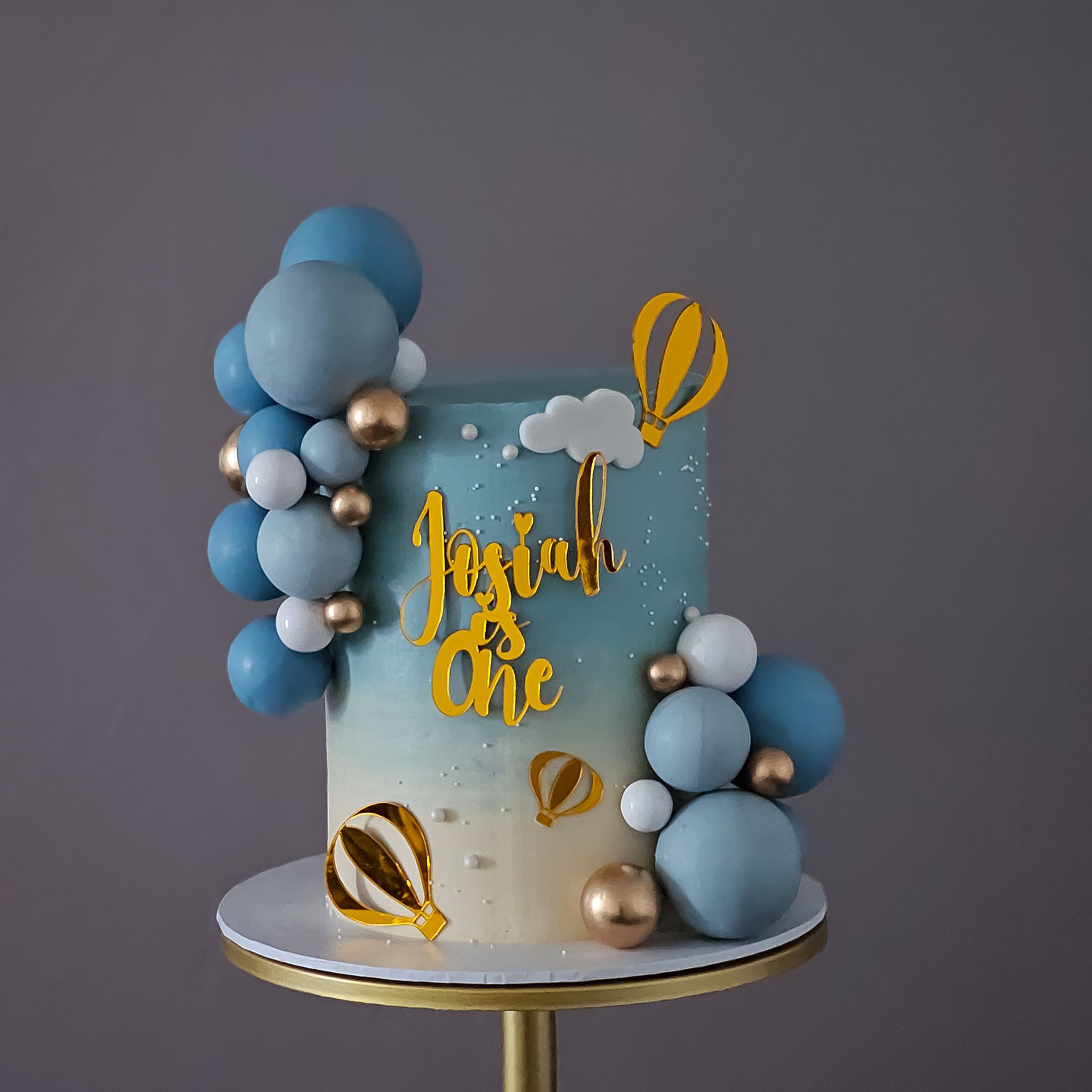 A birthday cake with a blue and white ombre design, decorated with blue and gold balloons, hot air balloon cutouts