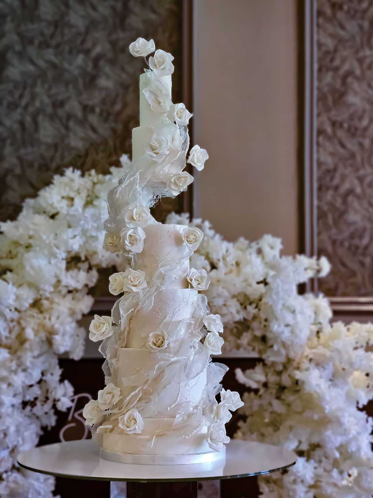 Elegant white wedding cake with floral decorations on a table surrounded by more white flowers.