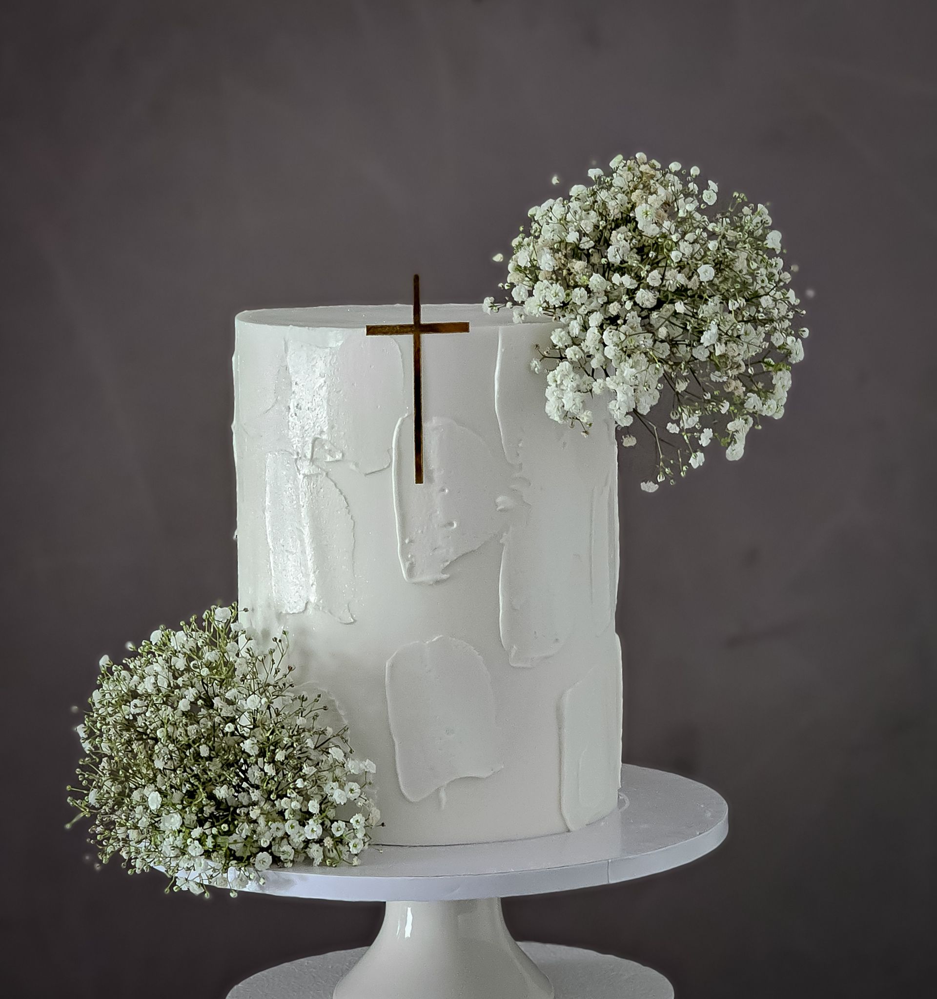White frosted cake on a pedestal, decorated with a gold cross and white baby's breath flowers.