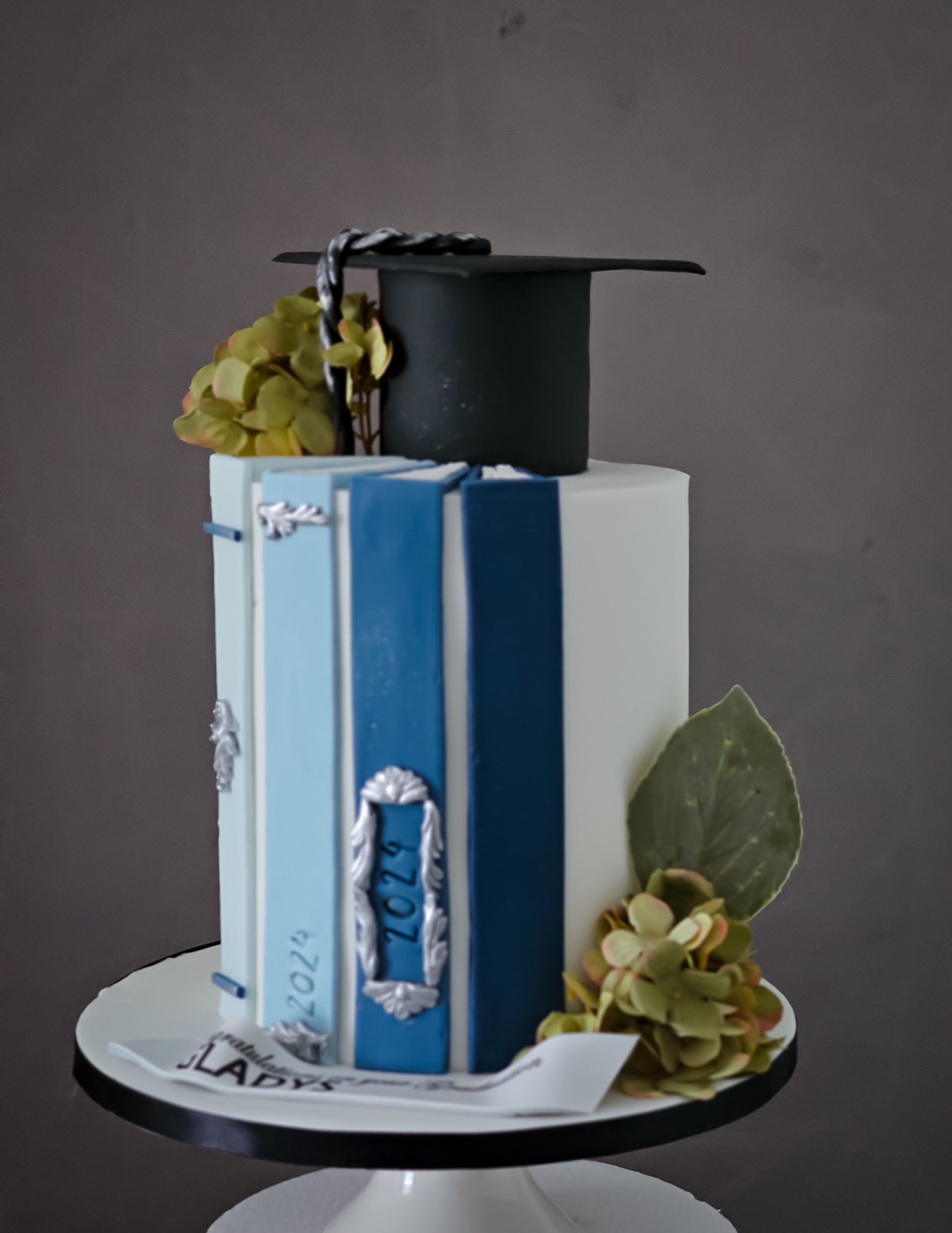 Graduation cake shaped like books, with a mortarboard on top and floral decorations, on a white cake stand.