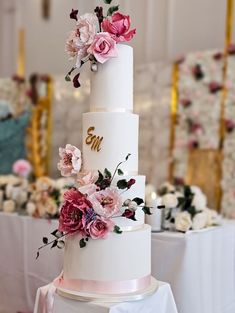 Four-tiered white wedding cake decorated with pink and burgundy flowers, gold initials, and a pink ribbon. 