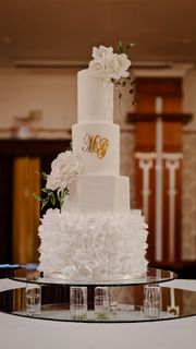 Four-tiered white wedding cake with floral decorations and gold initials, on a mirrored stand.