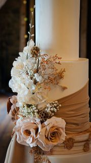 Three-tiered wedding cake decorated with ivory fondant, draped burlap, and floral arrangements of white and blush roses.
