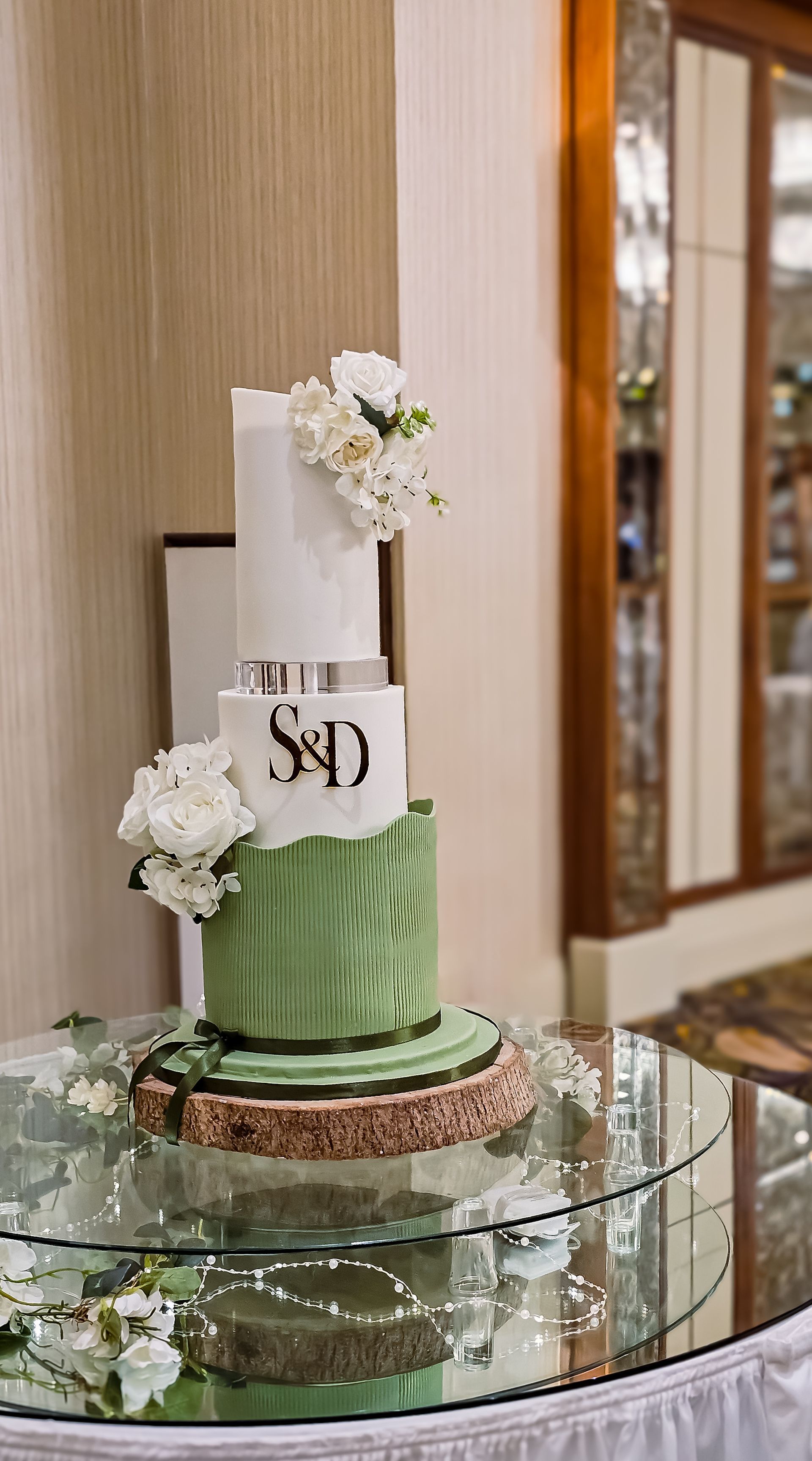 Three-tiered wedding cake with white and green layers, decorated with flowers and initials, on a mirrored table.