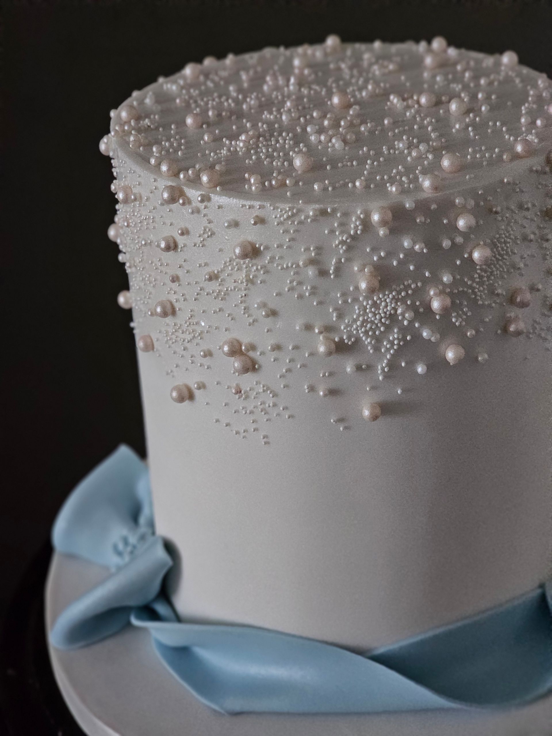 White cake decorated with edible pearls and sparkling sprinkles, tied with a blue ribbon.