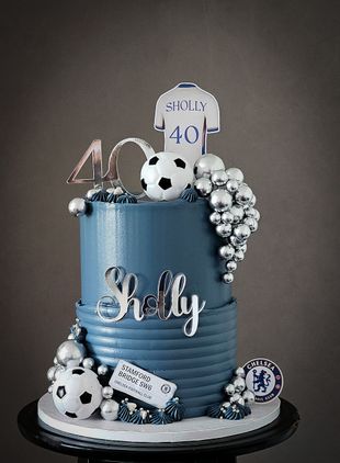 Two-tiered blue Chelsea-themed birthday cake, topped with a jersey, soccer balls, and silver accents.