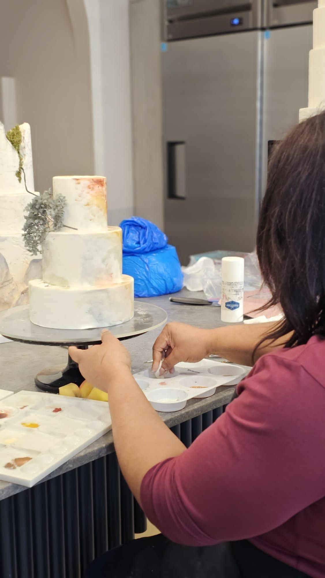 Person decorating a three-tiered cake with muted colors and foliage. They are working at a counter in a professional kitchen.