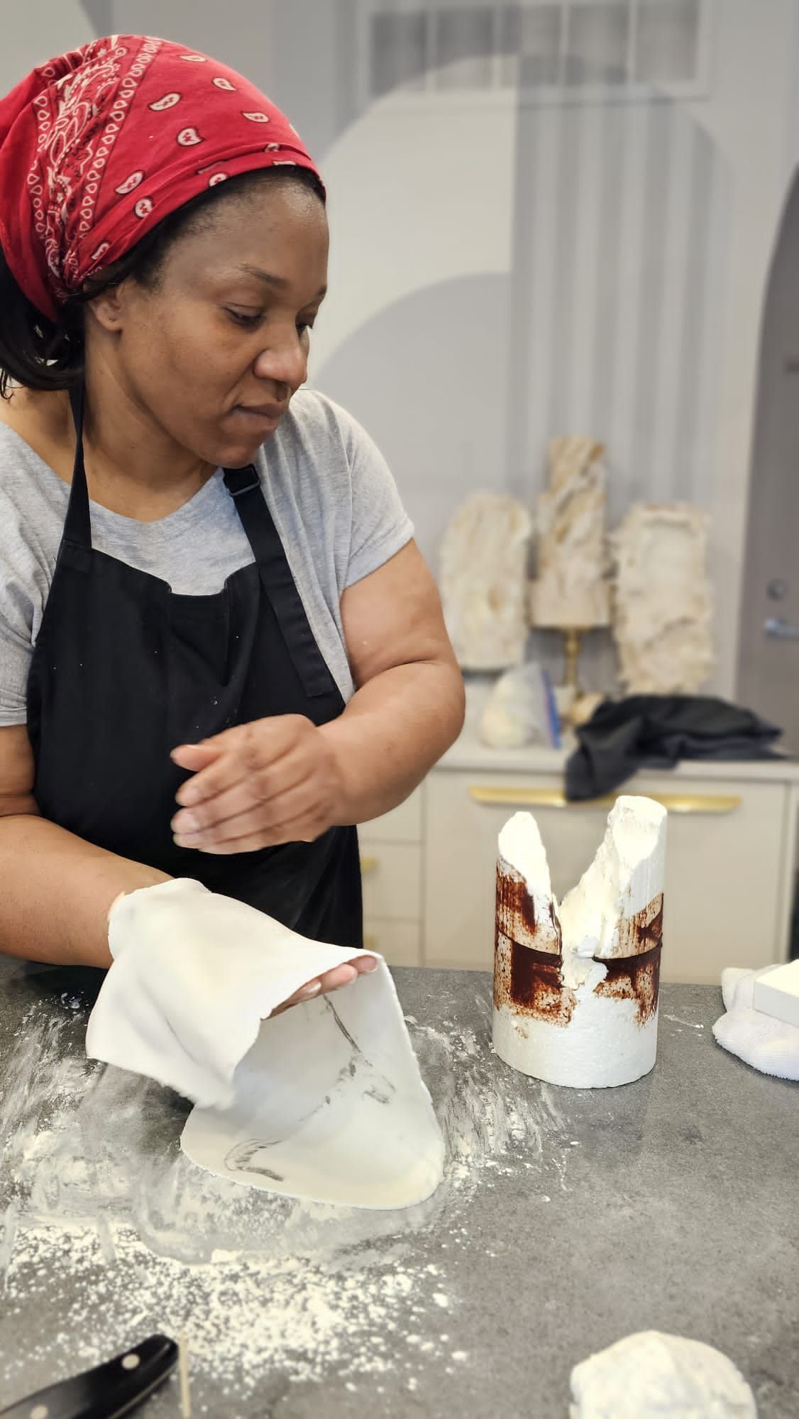 Woman in a headscarf and apron kneading dough on a floured countertop. Sculptures and baking supplies are in the background.