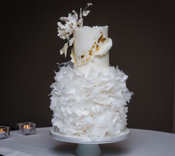Two-tiered white cake with ruffled bottom tier, smooth top tier with gold accents, and sugar flower decorations.