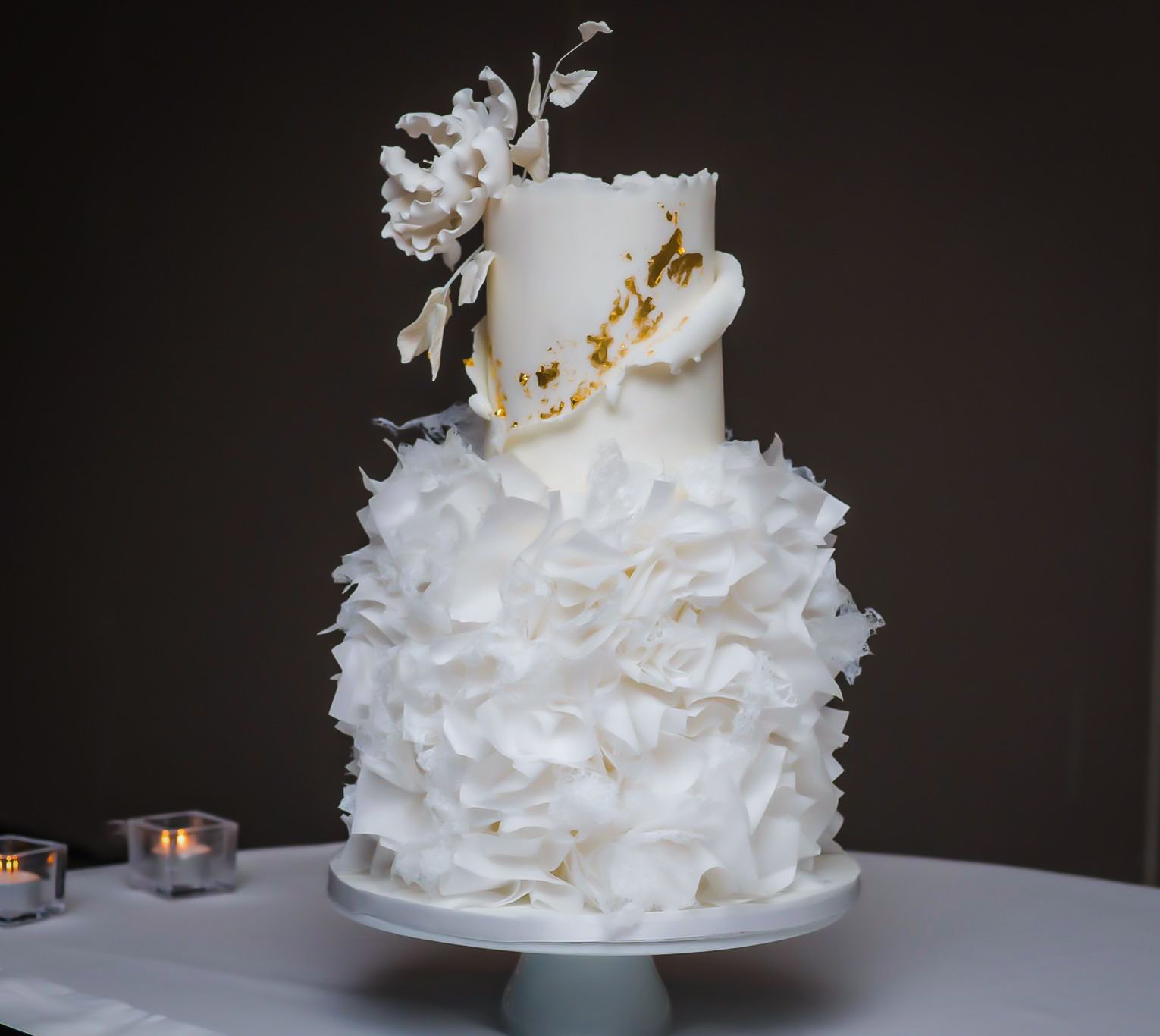 White wedding cake with ruffle-like skirt and flower decorations on a cake stand, lit by candlelight.