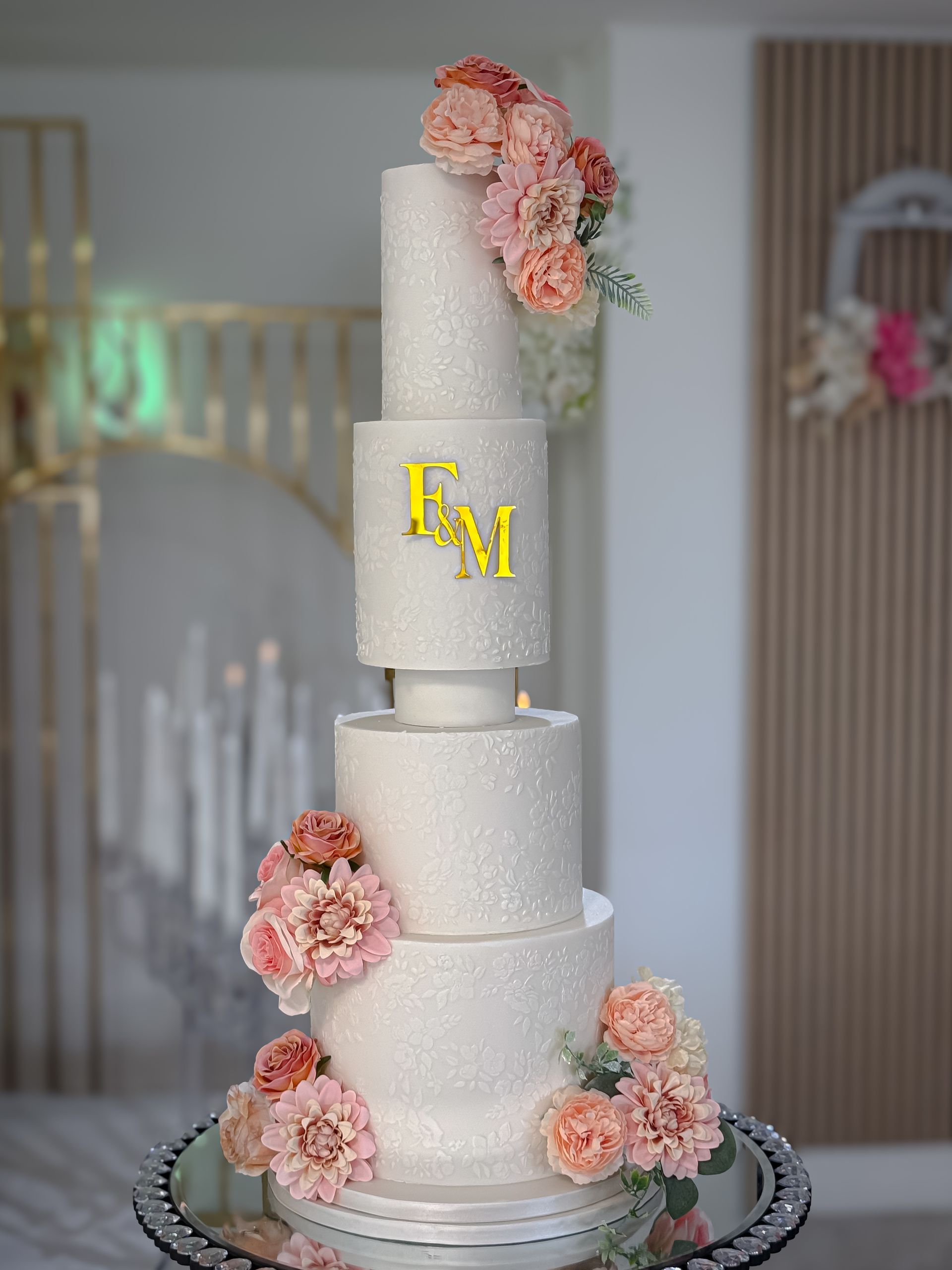 Five-tiered white glitter wedding cake with pink flowers and gold initials on a mirrored stand.