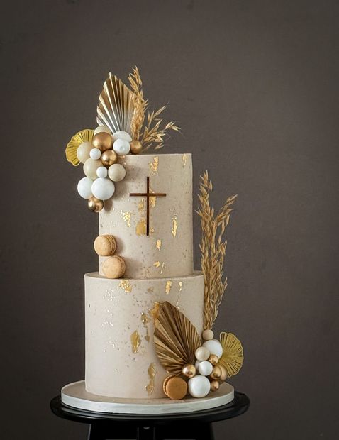 Two-tiered cake decorated with gold accents, a cross, and balloon-like spheres, set against a dark background.