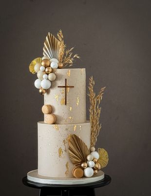 Two-tiered beige cake with gold accents, a cross, and decorative gold and white spheres and leaves.