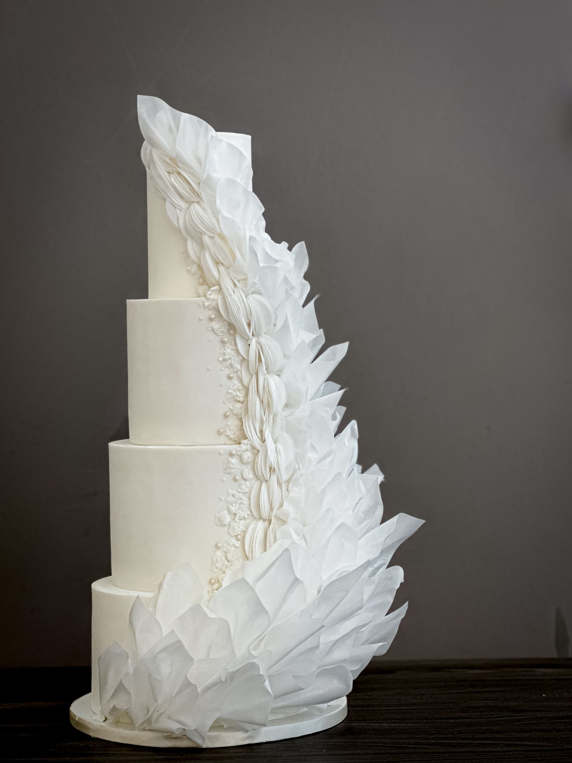 Four-tiered white wedding cake with sculpted sugar art cascading down the side, resembling ice crystals or fabric folds.