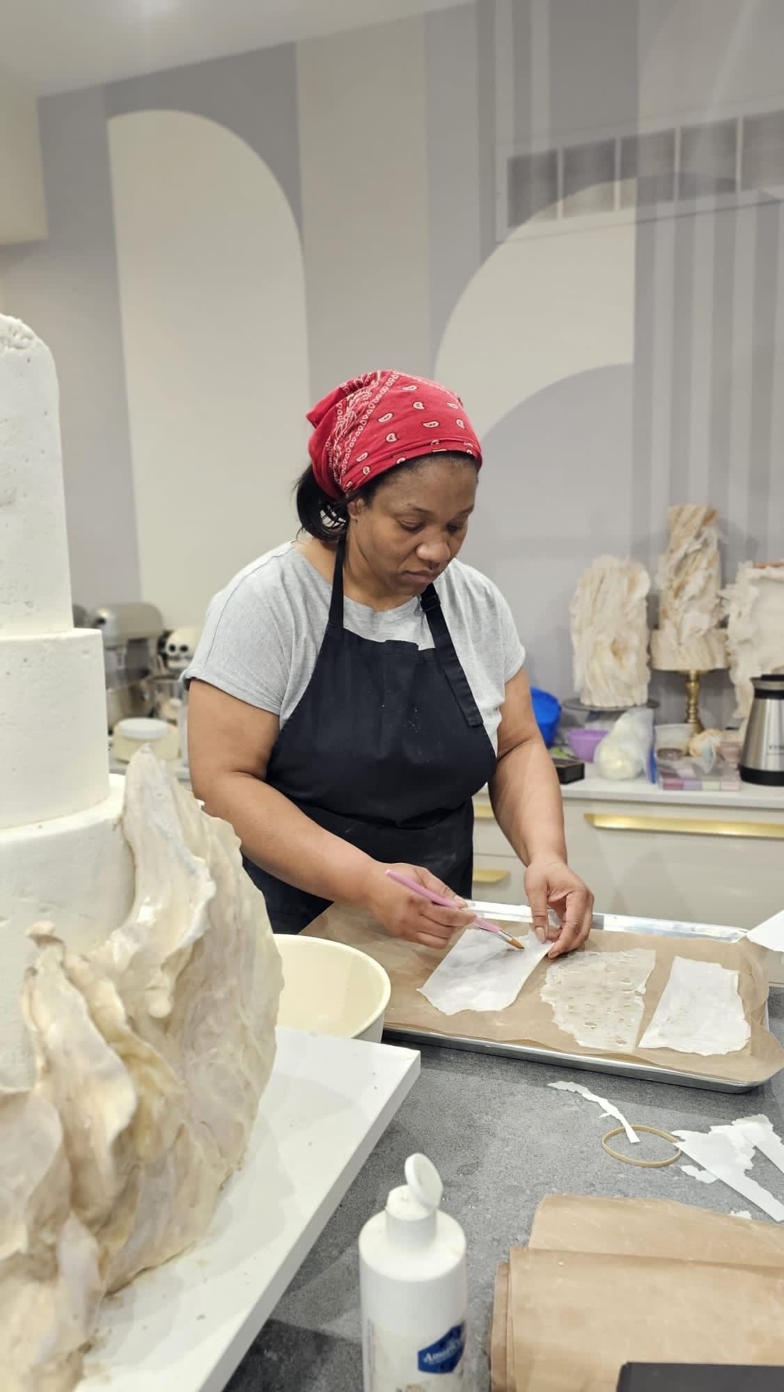 A woman in a red bandana and apron crafts cake decorations in a bright kitchen.