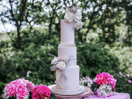 Four-tiered white wedding cake decorated with flowers, set outdoors with pink peonies in the foreground.