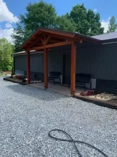 Covered porch with wooden posts beside a dark gray building, overlooking a gravel yard and trees.