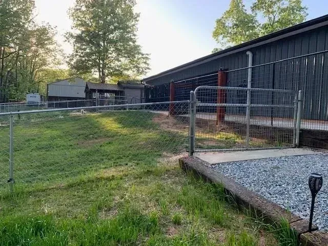 Chain-link fenced yard beside dark barn-like buildings with grass and a concrete walkway at sunset