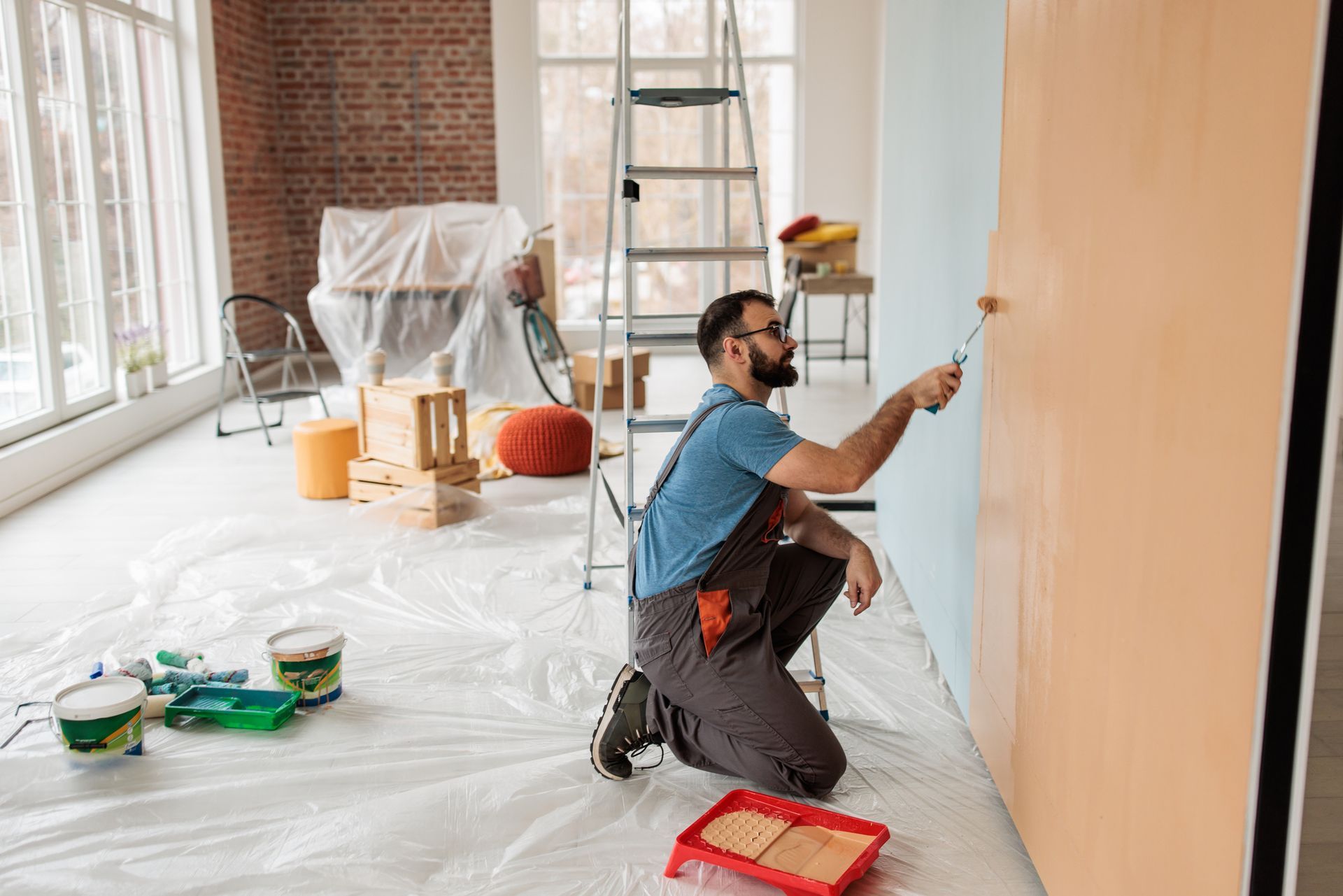 Person kneeling while painting a wall with a roller in a bright, prepped living space. Person kneeling while painting a wall with a roller in a bright, prepped living space.