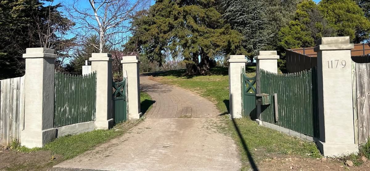 Open gate with green fence and stone pillars leading to a dirt path, trees in the background.
