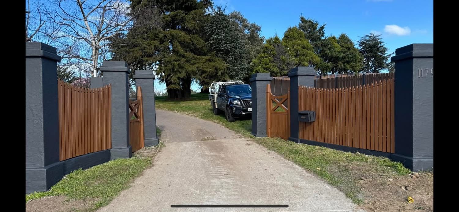 A driveway leading toward a house. The entrance has a wooden fence and black pillars. A vehicle is parked in front of the house.