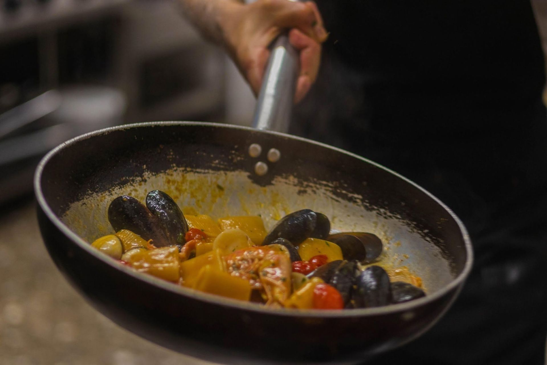 Una mano mescola cozze e verdure in una padella nera sul fornello.