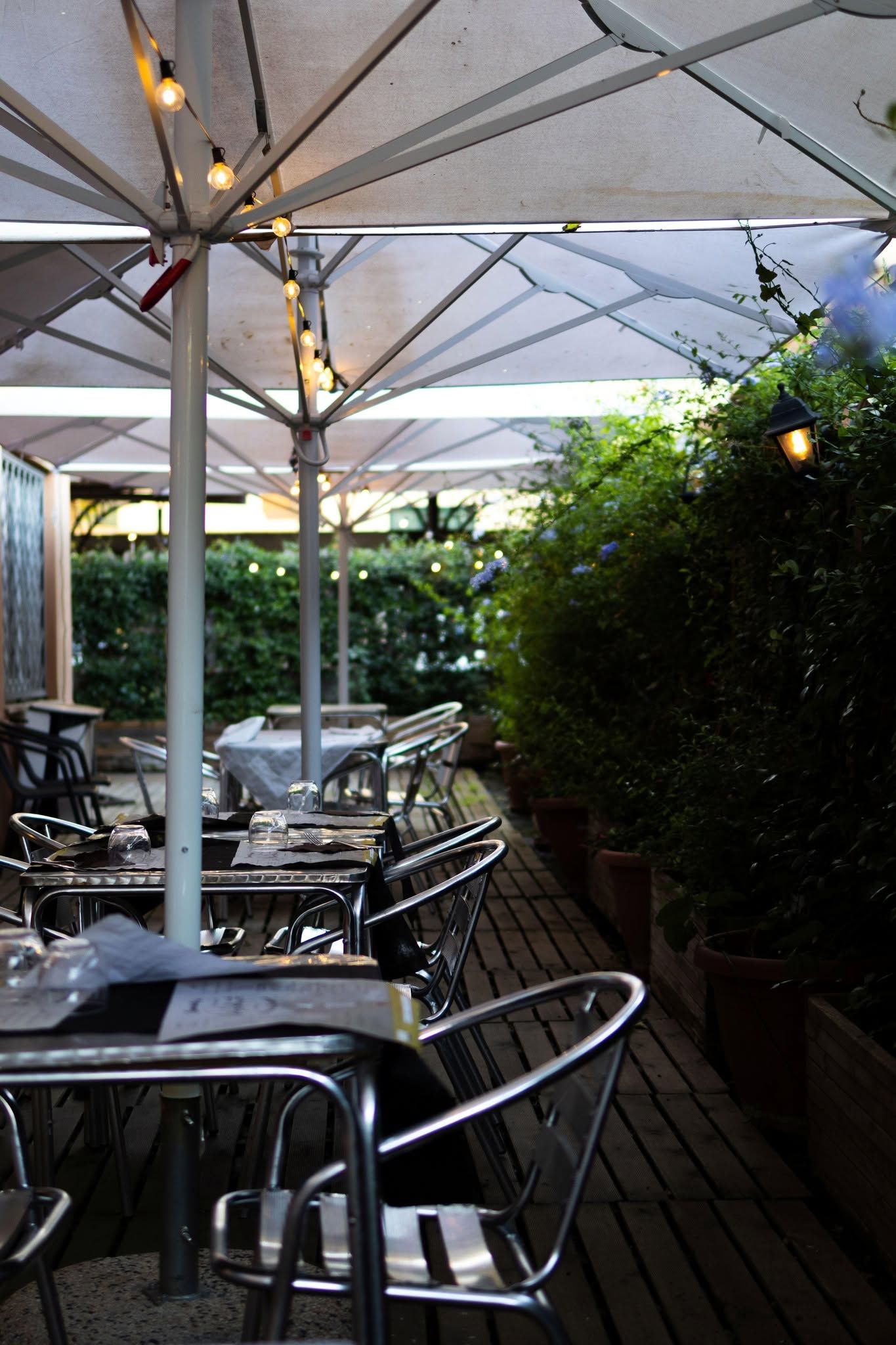 Tavolini all'aperto del caffè sotto grandi ombrelloni bianchi, accanto a una siepe e a una terrazza in legno.