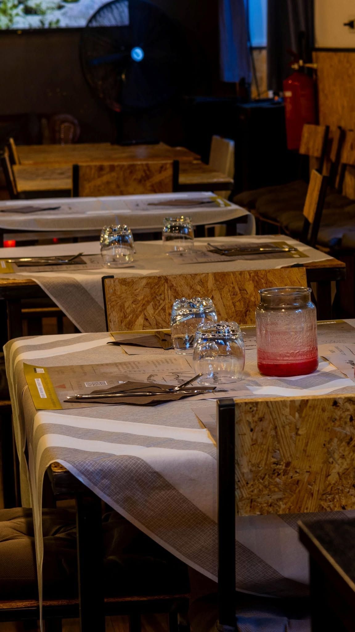 Tavoli di ristorante scarsamente illuminati con tovaglie bianche, bicchieri e una bevanda rosa su una panca a scacchi.