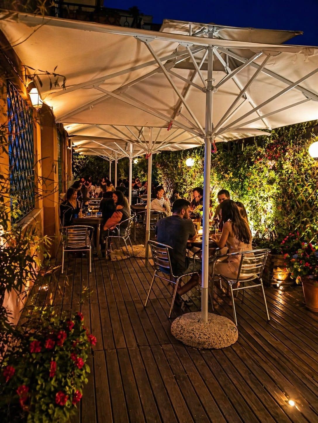 Cena all'aperto in terrazza di sera, con persone sedute sotto ombrelloni bianchi, luci soffuse e piante in vaso.