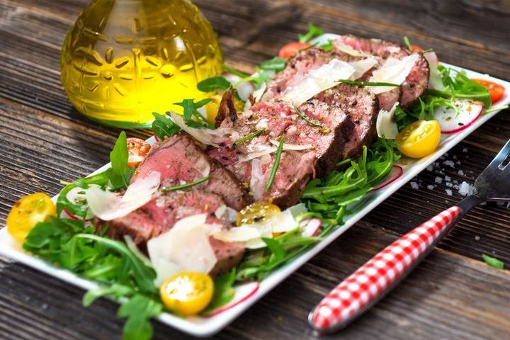 Fettine di bistecca servite con verdure, scaglie di formaggio e pomodorini su un tavolo di legno rustico.