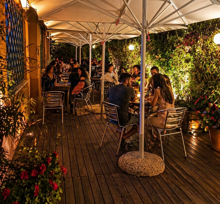 Di sera, il patio esterno di un caffè si affaccia sulla strada, con persone sedute ai tavoli sotto ombrelloni bianchi e luci soffuse dai toni caldi.