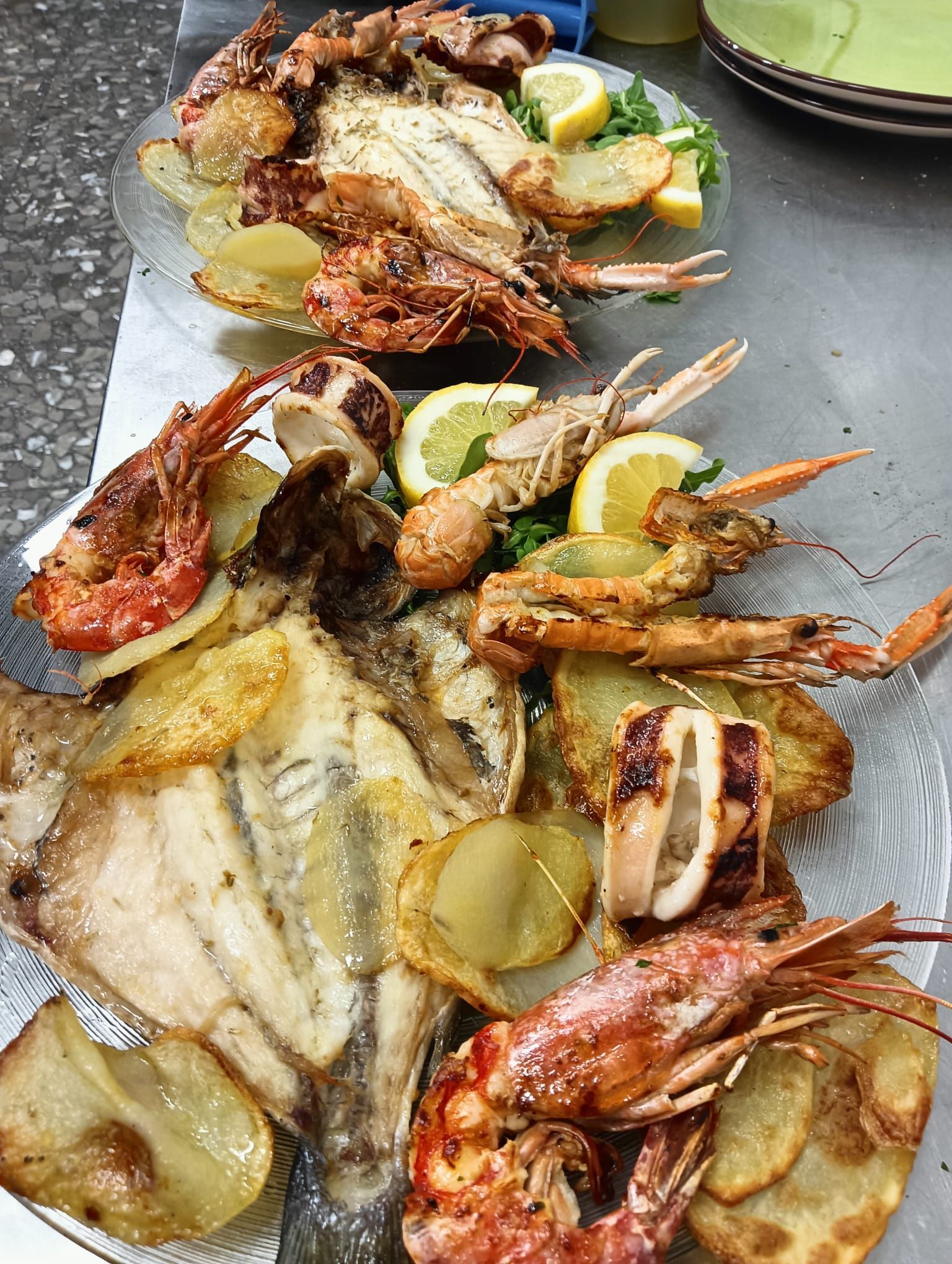 Piatto di frutti di mare grigliati con gamberi, pesce, fette di limone e patate su un vassoio scuro.