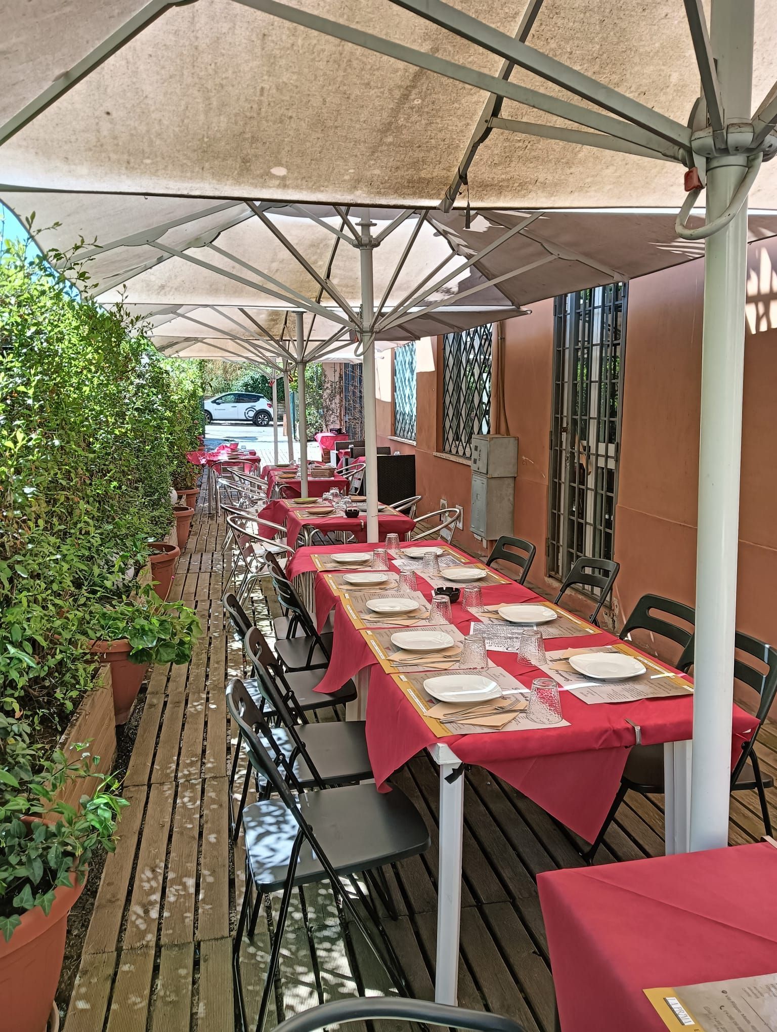 Dehor esterno del ristorante con tovaglie rosse, panchine in legno e ombrelloni beige lungo uno stretto passaggio.
