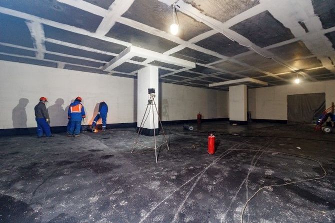 Workers in blue suits inspect a large unfinished room with exposed ceiling and construction tools.
