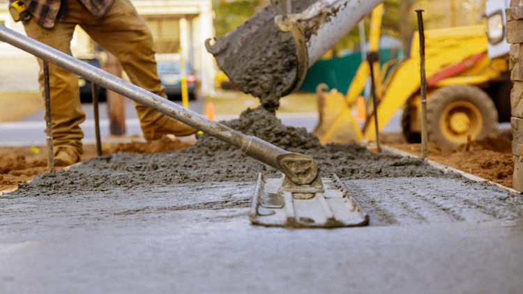Construction worker pouring concrete from a chute and leveling it with a screed tool.