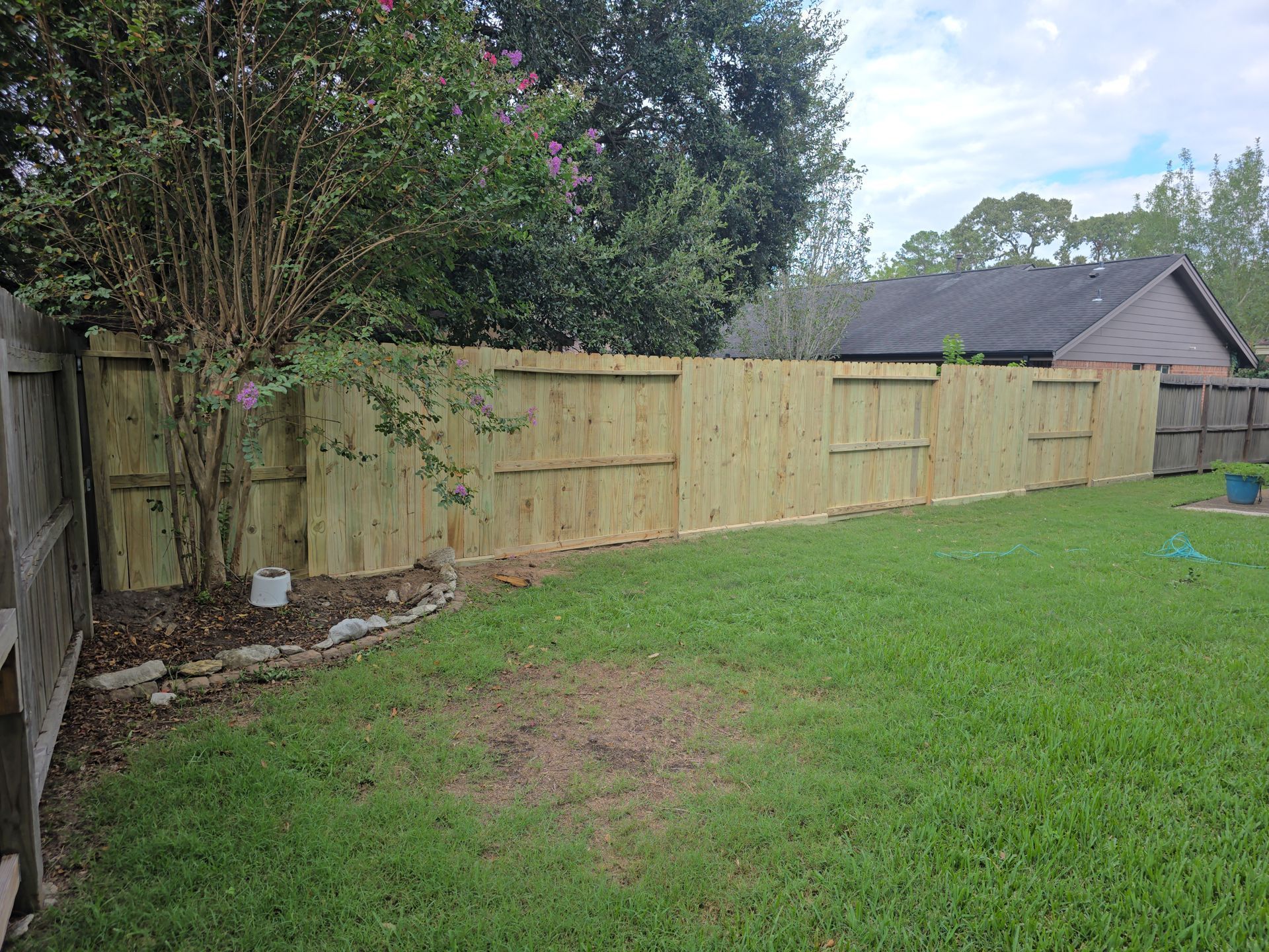 Backyard with green lawn and a long wooden privacy fence, trees and a house in the background