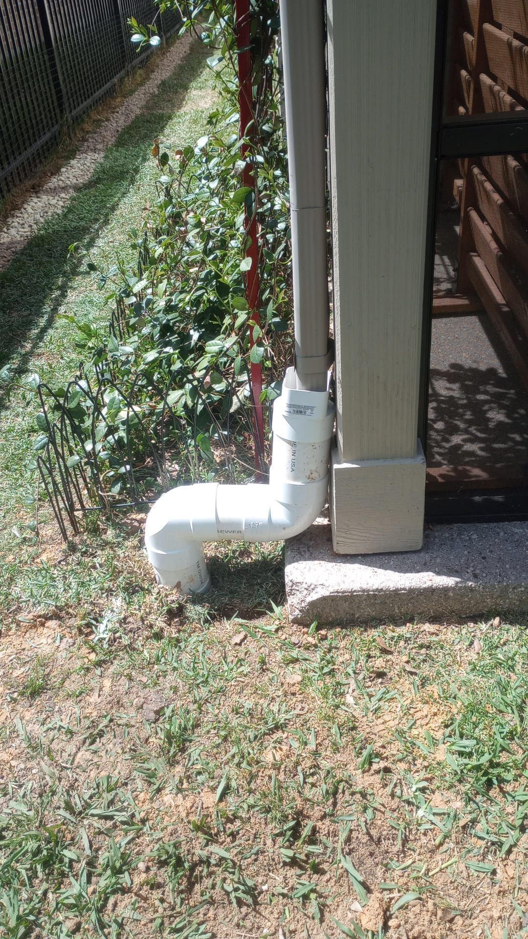 White PVC downspout elbow beside a house corner, draining near a grassy lawn and fence.