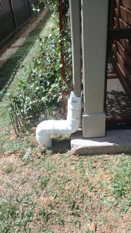 White PVC drainpipe beside a house corner, with a yard and fence in the background.