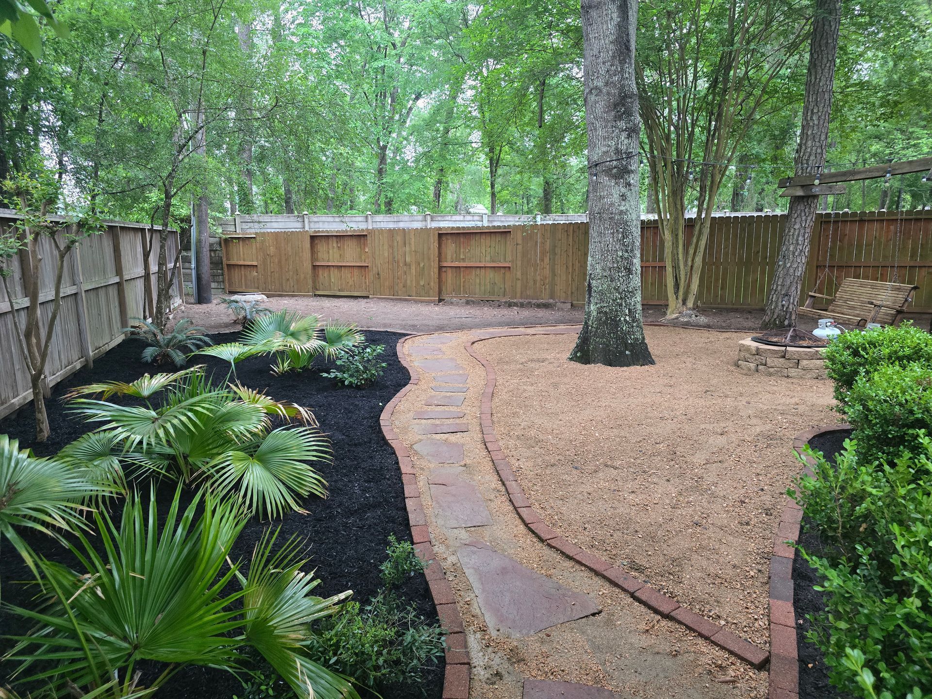 Curved stone path in a wooded backyard with ferns, mulch, trees, and a wooden fence