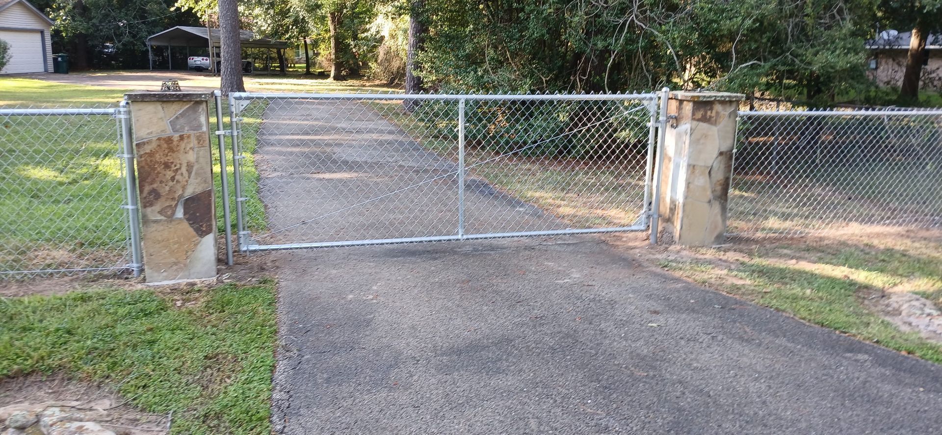 Chain-link driveway gate between stone pillars across a paved path, with trees and grass beyond