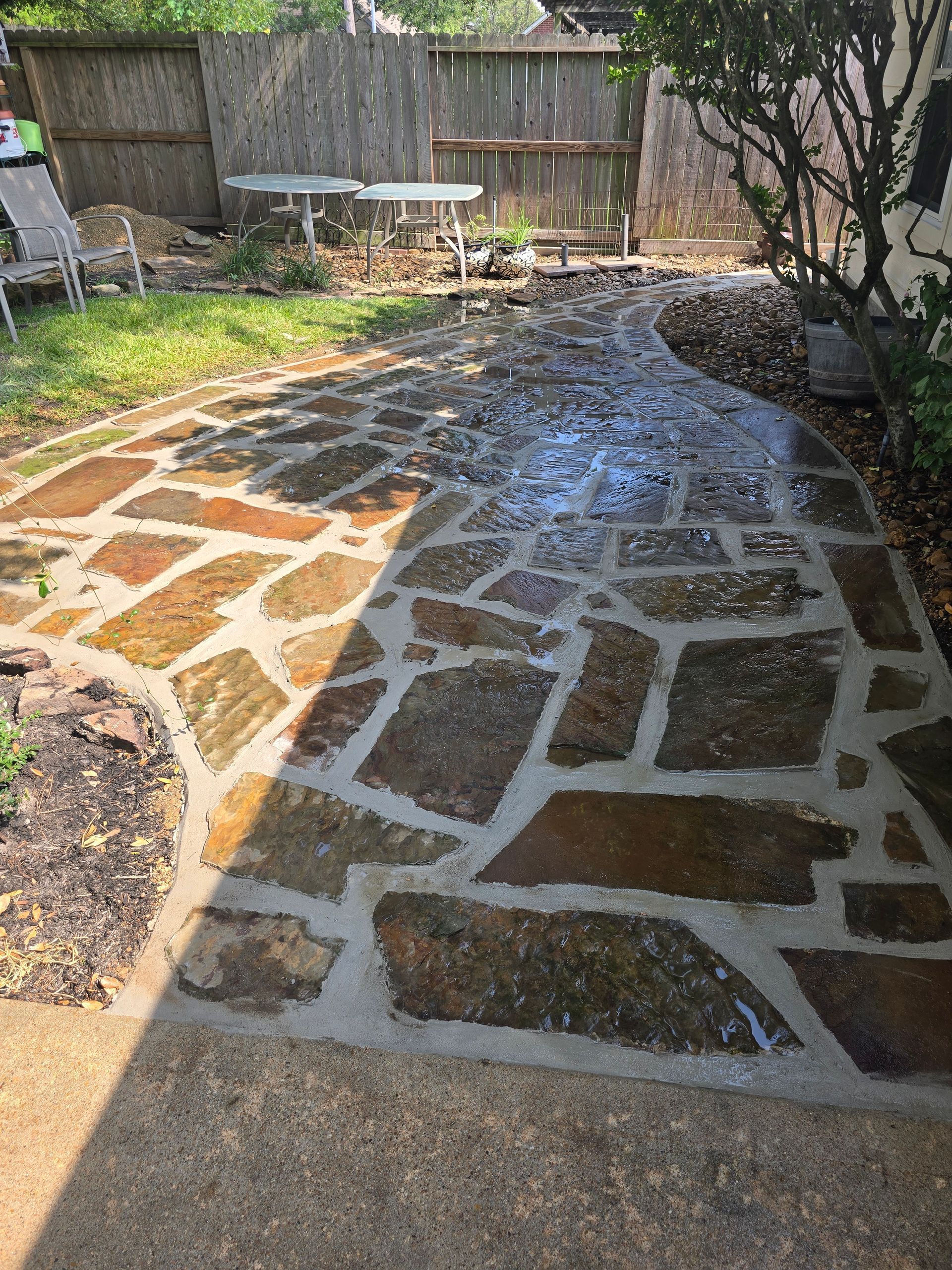 Stone backyard patio path with wet patches, bordered by grass and a wooden fence.