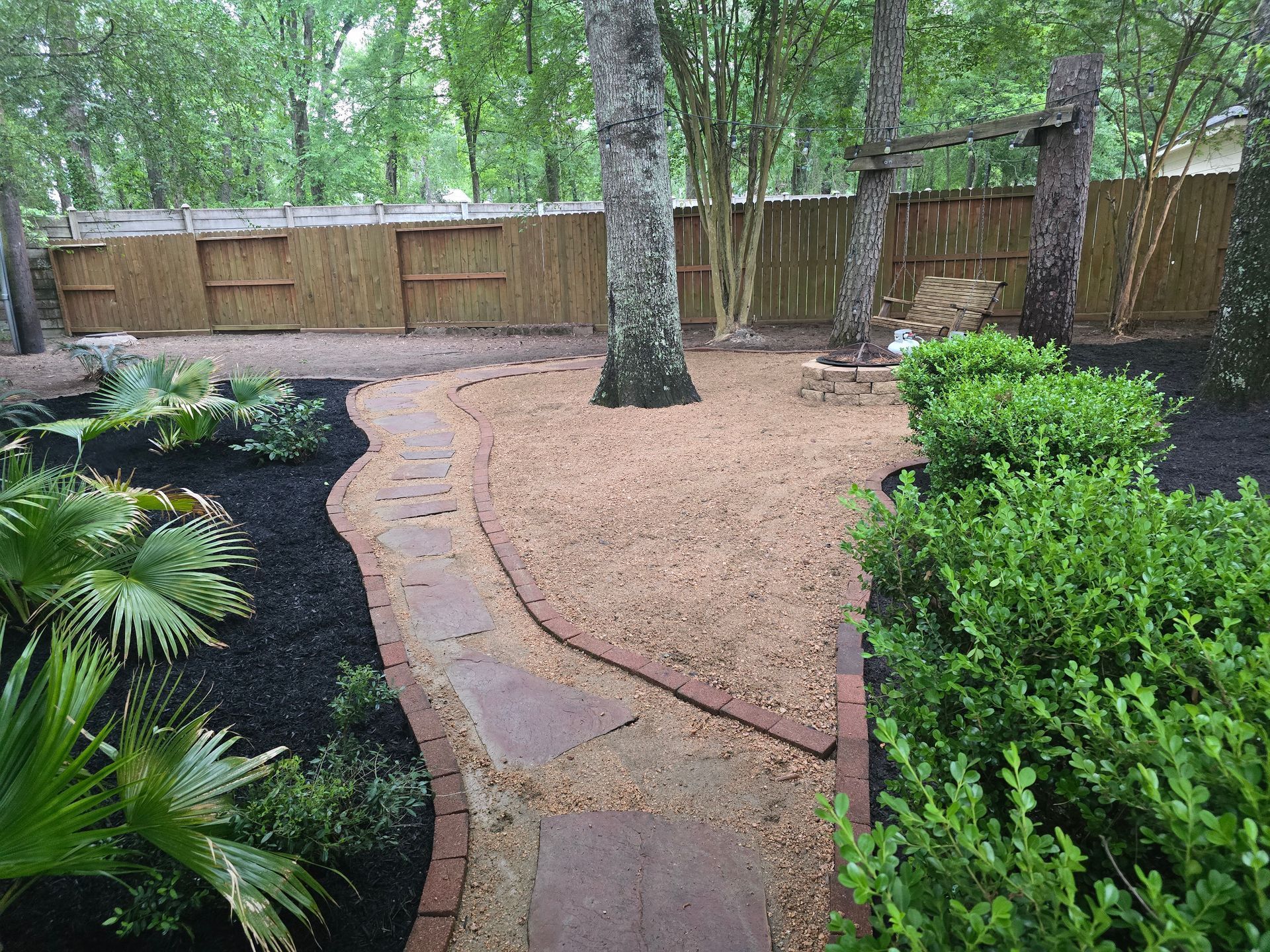 Curved stone path through a mulched garden with shrubs, palm, and trees beside a wooden fence