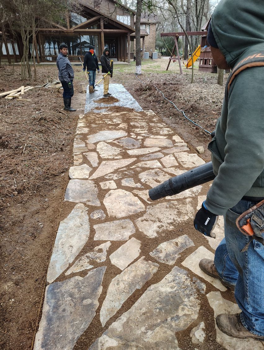 Stone walkway under construction in a wooded yard, with workers and tools visible along the path.