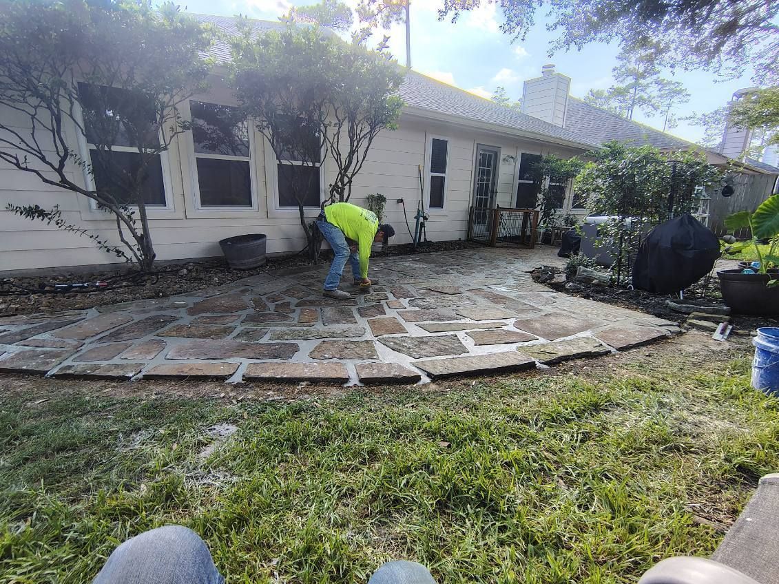 Workers repairing a stone patio beside a white house, with tools and a hose in the yard