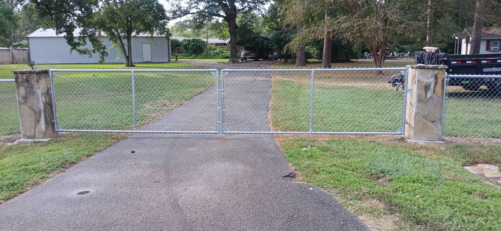 Gravel driveway leading to a closed chain-link gate between concrete posts, with a grassy yard beyond.
