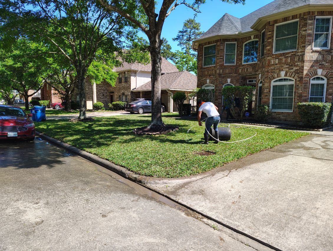 Apartment complex with a worker trimming grass beside a driveway and parked car on a sunny day