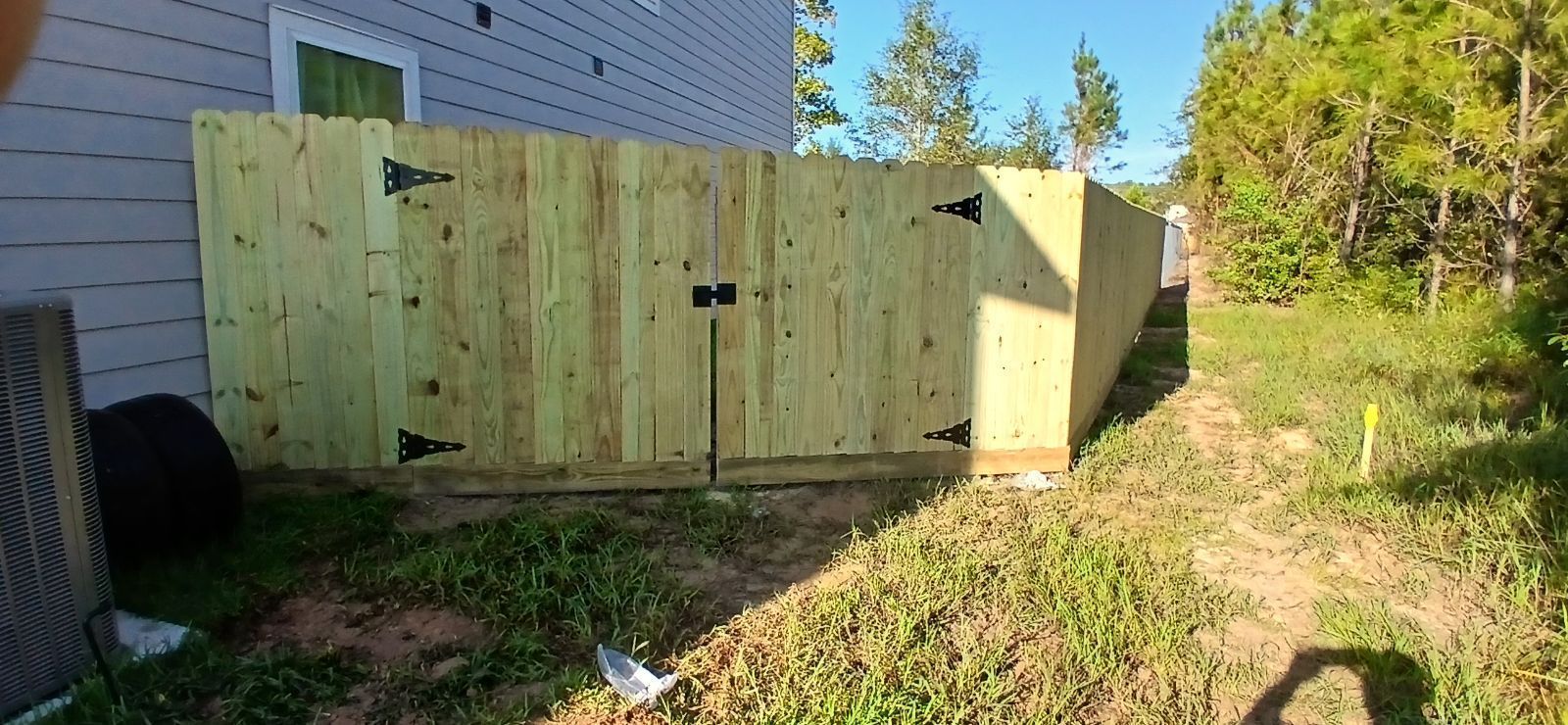 Wooden fence gate beside a house in a grassy yard, with trees in the background.
